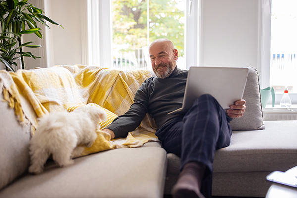 Older man on couch with dog and laptop on his lap